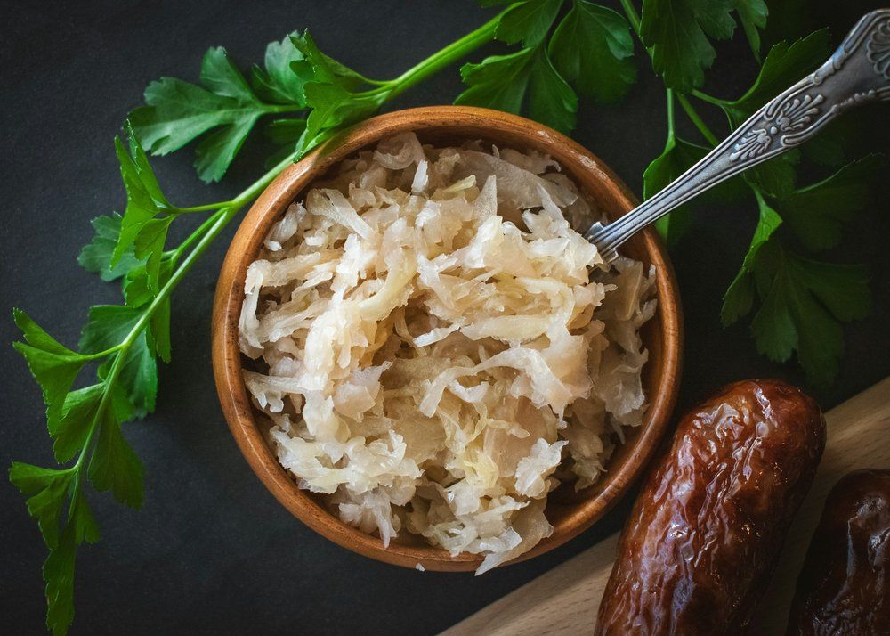 A wooden bowl of sauerkraut with a spoon, fresh parsley, and part of a grilled sausage.